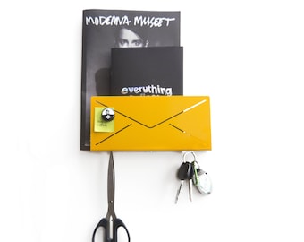 May include: A yellow metal envelope wall organizer with a black magazine and a black book with the text "everything" on the front. There are keys hanging from the bottom of the organizer and a pair of scissors hanging below. A small yellow sticky note with the text "buy coffee!" is attached to the organizer.