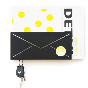 May include: Black metal envelope-shaped wall mounted key holder with a yellow circle sticker that says "SIKSA DESIGN" and a set of keys hanging from it. The key holder is holding a magazine with the text "DET Review of Architecture and Construction English Edition" on the cover.