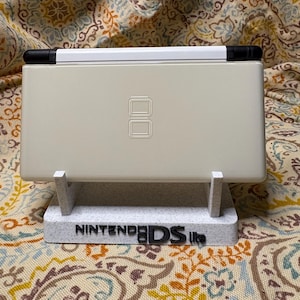 以下が含まれることがあります： クリーム色のニンテンドーDS Lite携帯ゲーム機が、カスタムスタンドに置かれています。スタンドはまだら模様のグレーで、「NINTENDO DS lite」の文字が黒で表示されています。コンソールは開いており、上部画面と黒いヒンジが見えています。