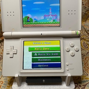 Peut inclure: Console de jeu portable Nintendo DS Lite blanche, ouverte pour afficher un jeu Mario. L'écran supérieur affiche une scène de jeu colorée, tandis que l'écran inférieur affiche un menu de sélection de jeu. La console repose sur un support blanc avec "NINTENDO DS lite" imprimé.