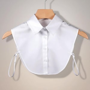 Pode incluir: Uma gola branca destacável e impecável com um design pontudo clássico. A gola apresenta uma frente abotoada e fitas delicadas. Está pendurada numa cabide de madeira contra um fundo neutro, mostrando as suas linhas limpas e o seu estilo versátil.