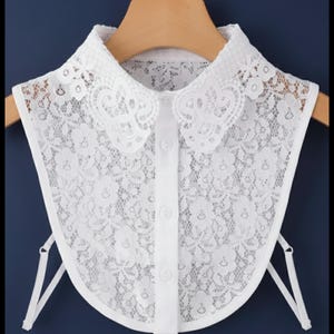 Pode incluir: Um colarinho removível branco feito de tecido de renda floral. O colarinho apresenta um design arredondado com bordas festonadas, uma carcela central vertical com três botões brancos e tiras elásticas finas e ajustáveis nos lados.