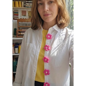 May include: A white button-down shirt layered over a yellow t-shirt, featuring a vertical row of six bright pink crocheted flower embellishments attached to the button placket, each centred with a small white button detail.