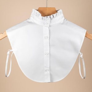 May include: A white detachable shirt collar with a ruffled high collar and a button-down front. The collar is hanging on a wooden hanger and has adjustable straps on each side.