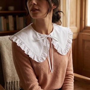 May include: A white, ruffled collar with a tie closure. The collar is worn over a peach-colored sweater. The ruffles add a decorative element to the collar. The background includes a bookshelf and wooden paneling.