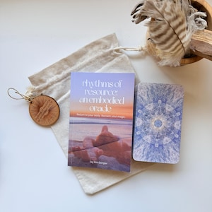 May include: A deck of oracle cards titled Rhythms of Resource: An Embodied Oracle by Kim Gengler, featuring a guidebook with a beach sunset cover, a deck of cards with blue mandala patterns, a small wooden charm, and a beige cotton drawstring pouch.
