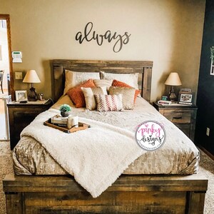 A large metal sign cut out to read the word always. The sign is painted black and mounted above a farmhouse looking headboard. The bed is made with decorative pillows and books. There is a pinky designs logo in the bottom right of the bed.