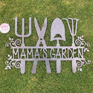 A metal gardening decor piece with 4 gardening tools, there is a line in between them for the wording. The sign reads Mamas Garden and is left raw metal. The sign is laid on green grass to show contrast. There is a pinky designs logo in the upper lef