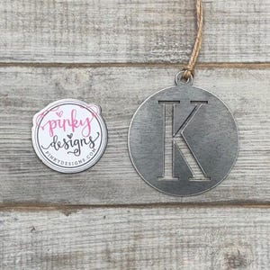May include: A metal ornament shaped like a circle with the letter K cut out of the center. The ornament is hanging from a twine string. A sticker with the text "pinky designs" is in the background.