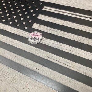 以下が含まれることがあります： 白木目調の背景に、黒い金属製のアメリカ国旗の壁飾り。旗は薄い金属のストリップで作られており、星形のカットアウトデザインが施されています。画像の左下隅に「pinky designs」の文字が見えます。