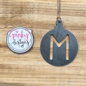 May include: A gray wooden ornament shaped like a Christmas tree bauble with the letter M cut out in the center. The ornament is hanging from a string.