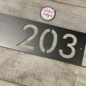 There is a horizontal address sign reading 203 and is left aligned on the plaque. The metal is left Raw Metal really show casing the details of the raw metal patina. It's laid on a light wood background with a pinky designs logo above. Another angle.