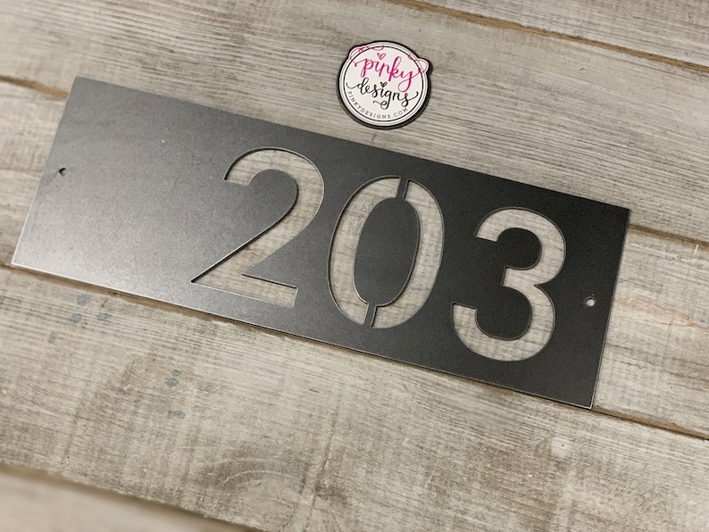 There is a horizontal address sign reading 203 and is left aligned on the plaque. The metal is left Raw Metal really show casing the details of the raw metal patina. It's laid on a light wood background with a pinky designs logo above. Another angle.