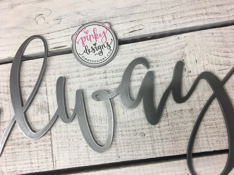 A metal always sign is laid on a light wood background. The metal is left raw metal to show the patina of the natural metal. There is a pinky designs logo located above the metal piece