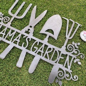 A metal gardening decor piece with 4 gardening tools, there is a line in between them for the wording. The sign reads Mamas Garden and is left raw metal. The sign is laid on green grass to show contrast. This is a repeated sign from another angle
