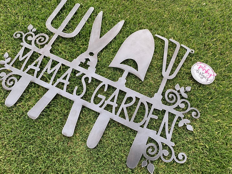 A metal gardening decor piece with 4 gardening tools, there is a line in between them for the wording. The sign reads Mamas Garden and is left raw metal. The sign is laid on green grass to show contrast. This is a repeated sign from another angle