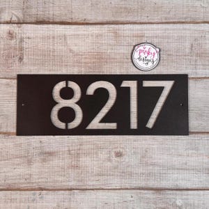 May include: A rectangular black metal house number sign featuring the cut-out digits 8217. The sign has two small circular mounting holes on the left and right sides. A small circular sticker with the text pinky designs is positioned above the sign.