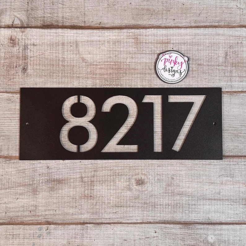May include: A rectangular black metal house number sign featuring the cut-out digits 8217. The sign has two small circular mounting holes on the left and right sides. A small circular sticker with the text pinky designs is positioned above the sign.