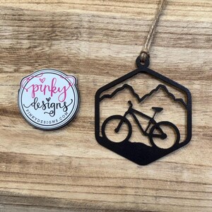 May include: Black metal ornament shaped like a hexagon with a mountain range and a mountain bike inside. The ornament has a twine string for hanging. The ornament is made by Pinky Designs.