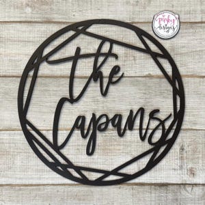 May include: Black wooden sign with a geometric frame and the text "The Capans" in a cursive font.