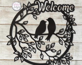 May include: Black metal wall decor featuring a circular design with the word "Welcome" at the top. Two birds are perched on a branch with leaves, creating a nature-inspired design.