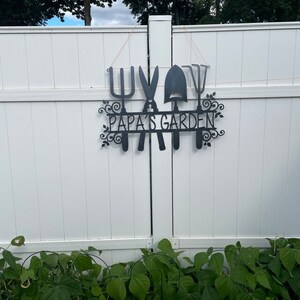 A metal gardening decor piece with 4 gardening tools, there is a line in between them for the wording. The sign reads Papas Garden and is left raw metal and mounted on a white wooden fence. Below the garden sign you can see vibrant greenery.