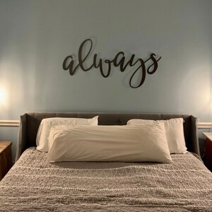 A large metal sign cut out to read the word always. The sign is painted black and mounted above a modern looking headboard. The bed is made with beautiful white pillows. There is lights above either side table on both sides of the bed.