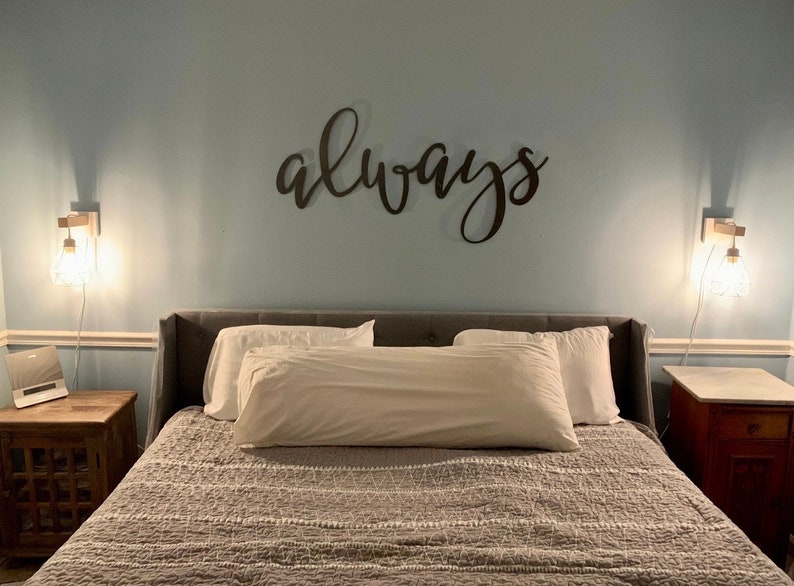 A large metal sign cut out to read the word always. The sign is painted black and mounted above a modern looking headboard. The bed is made with beautiful white pillows. There is lights above either side table on both sides of the bed.
