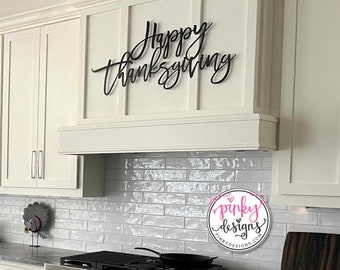 May include: Black metal Happy Thanksgiving sign hanging above a white kitchen cabinet. The sign is in a cursive font. A white tile backsplash is behind the sign. A black frying pan is on a stainless steel stovetop. A white and pink logo for Pinky Designs is in the lower right corner of the image.