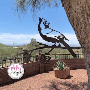 May include: A black metal cardinal bird sculpture perched on a branch with a pine cone. The sculpture is mounted on a tree trunk and is in front of a brick wall and a green plant.