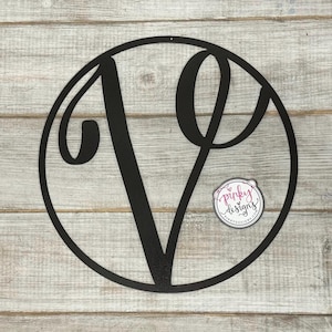 May include: Black metal monogram letter V in a circle on a light wood background. The letter is in a script font. The circle is about 12 inches in diameter.