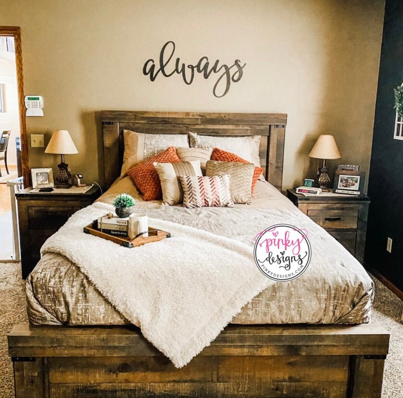 A large metal sign cut out to read the word always. The sign is painted black and mounted above a farmhouse looking headboard. The bed is made with decorative pillows and books. There is a pinky designs logo in the bottom right of the bed.