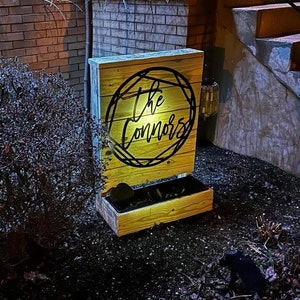 A metal last name sign reading The Connors. The sign is mounted on the back of a wooden planter. There is a spotlight on the Connors sign making it easy to read even though the photo is taken at night.