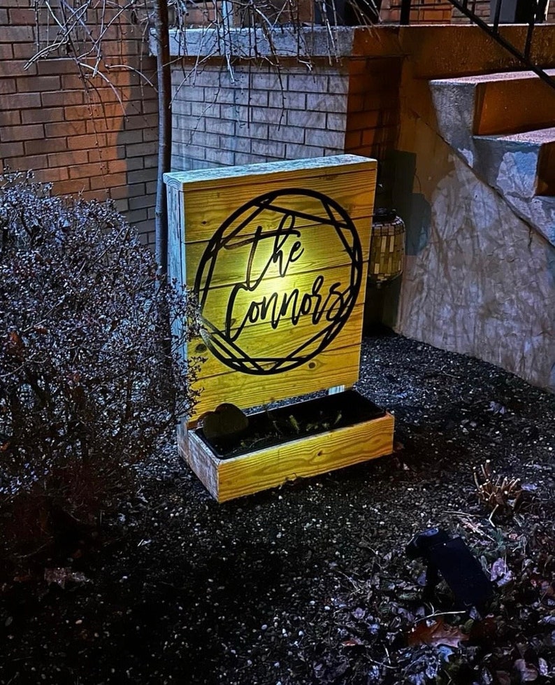A metal last name sign reading The Connors. The sign is mounted on the back of a wooden planter. There is a spotlight on the Connors sign making it easy to read even though the photo is taken at night.