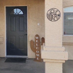 May include: A wooden cactus sign with the word "WELCOME" painted in white letters. The sign is standing on a porch with a black door and a metal cactus design on the wall.