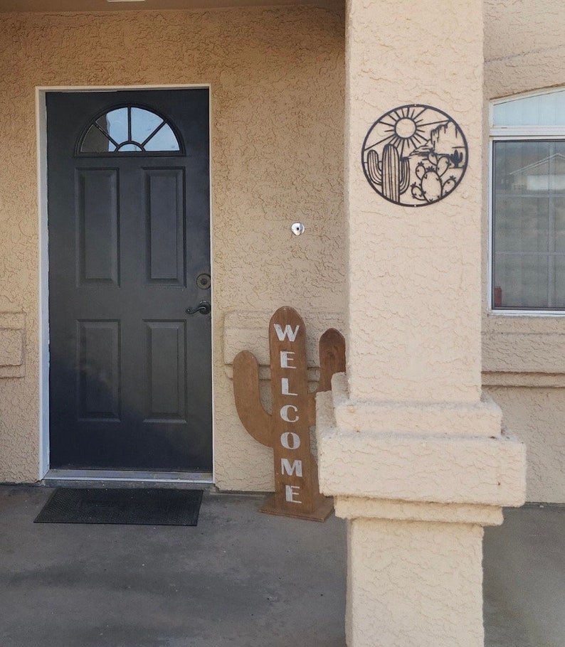 May include: A wooden cactus sign with the word "WELCOME" painted in white letters. The sign is standing on a porch with a black door and a metal cactus design on the wall.