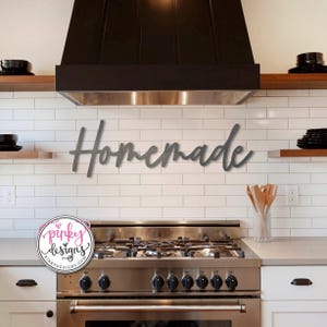 May include: A gray metal sign with the word "Homemade" in a cursive font, mounted on a white tiled wall above a stainless steel stove in a kitchen.