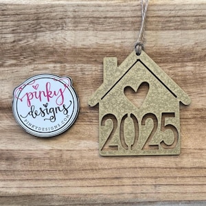 May include: A gold-colored wooden ornament shaped like a house with a heart cutout and the year 2025 inside. The ornament has a string for hanging.