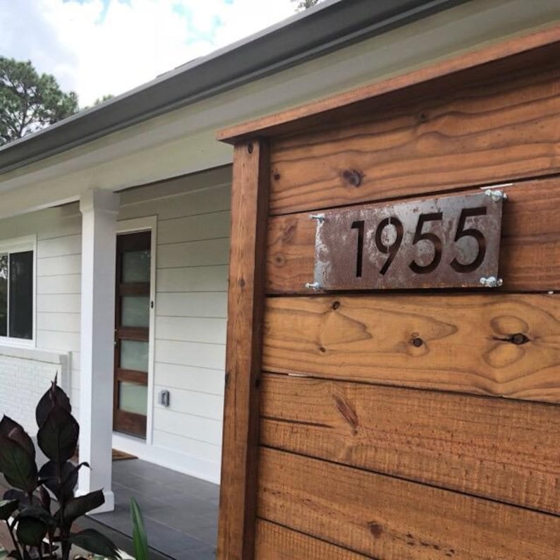 A modern rectangle sign with the numbers 1955 cut out of it. The sign is mounted on a wooden fence in front of the home. The metal was left raw metal and left to rust. The rusting patina gives a unique look to the house number sign.
