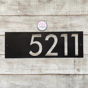May include: A rectangular black metal house number sign featuring the cut-out digits 5211. The sign has a small circular mounting hole on the far left and right edges. Above the sign is a circular logo sticker with the text pinky designs.
