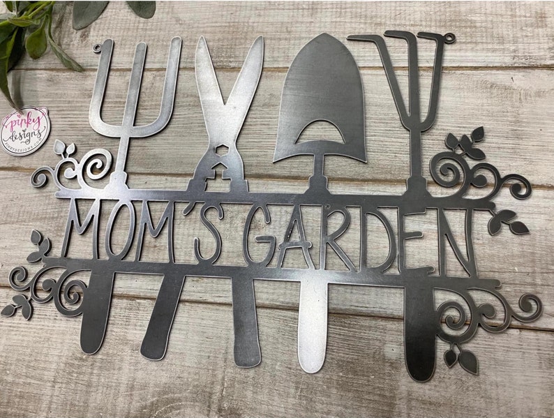 A metal garden home decor piece with the words Moms Garden cut out of it. The design has four gardening tools with a line in the middle for the saying. The metal is laid on a light wood background with a pinkydesigns logo to the left of the sign.