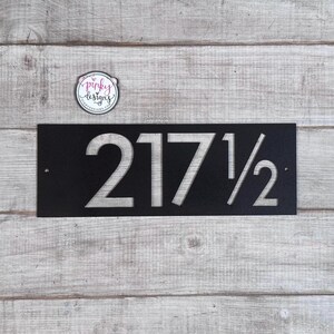 May include: A rectangular black metal house number sign featuring the cut-out numbers 217 1/2. The sign has two small circular mounting holes on the left and right sides. A circular logo sticker with the text pinky designs is positioned above.