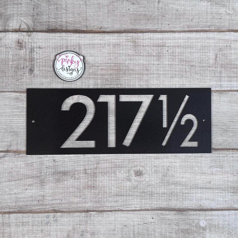 May include: A rectangular black metal house number sign featuring the cut-out numbers 217 1/2. The sign has two small circular mounting holes on the left and right sides. A circular logo sticker with the text pinky designs is positioned above.