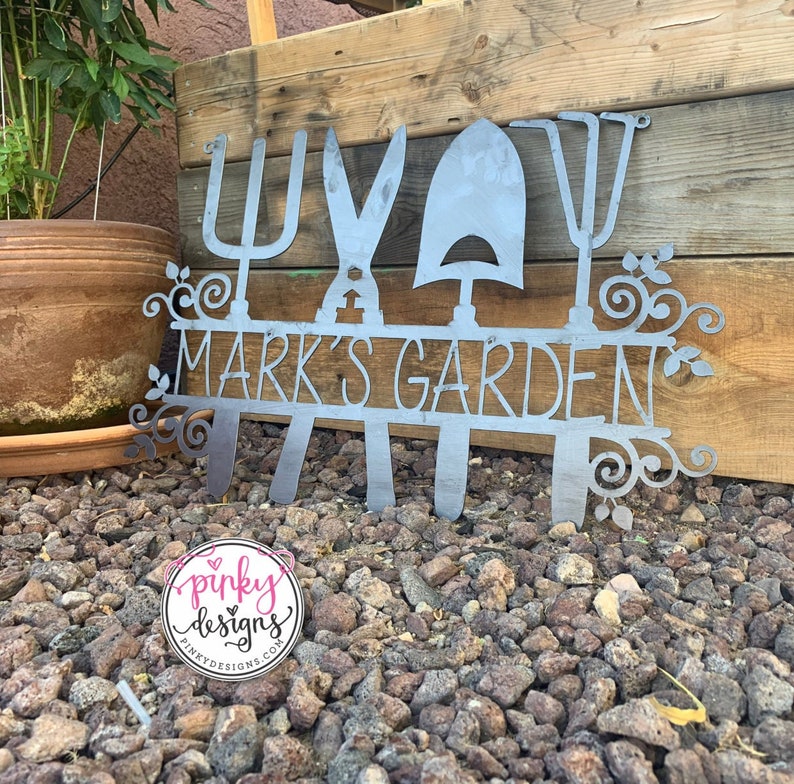 A metal gardening decor piece with 4 gardening tools, there is a line in between them for the wording. The sign reads Marks Garden and is left raw metal. The sign is sitting in rocks leaning against a raised garden bed. Pinky Designs logo can be seen