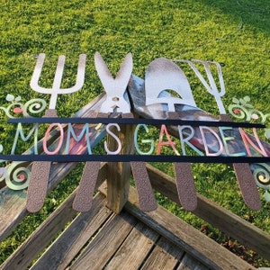 A metal gardening decor piece with 4 gardening tools, there is a line in between them for the wording. The sign reads Moms garden. The customer did a DIY project on their garden sign. The wording looks tie-dye while the tools are silver with dirt.