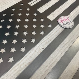 以下が含まれることがあります： 星とストライプのカットアウトデザインが施された金属製のアメリカ国旗。旗は白く塗られた木製の背景の上に置かれています。画像の右上隅にあるステッカーに「pinky designs」というテキストが表示されています。