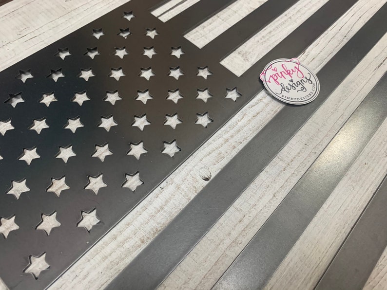 以下が含まれることがあります： 星とストライプのカットアウトデザインが施された金属製のアメリカ国旗。旗は白く塗られた木製の背景の上に置かれています。画像の右上隅にあるステッカーに「pinky designs」というテキストが表示されています。
