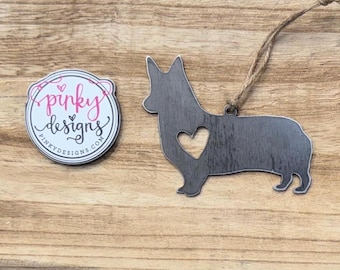 May include: A metal ornament shaped like a corgi with a heart cut out of its chest. The ornament is painted a dark grey colour and has a rustic finish. The ornament is hanging from a twine string. The ornament is accompanied by a round sticker with the text "pinky designs" and a website address.