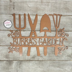 A metal garden home decor piece with the words Bubbas Garden cut out of it. The design has four gardening tools with a line in the middle for the saying. The metal is laid on a light wood background and is painted a copper color.