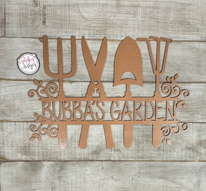 A metal garden home decor piece with the words Bubbas Garden cut out of it. The design has four gardening tools with a line in the middle for the saying. The metal is laid on a light wood background and is painted a copper color.